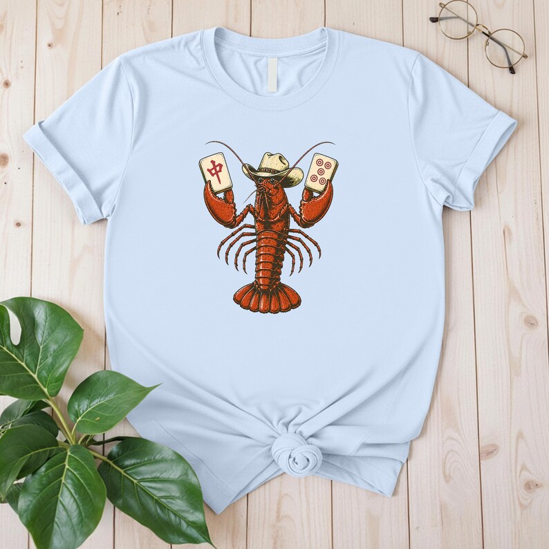 May include: A light blue t-shirt featuring a graphic illustration of a red lobster wearing a cowboy hat. The lobster is holding two mahjong tiles, one with a red Chinese character and the other with five red circles, centered on the chest.
