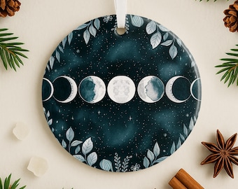 May include: A round ceramic ornament featuring a watercolor depiction of the phases of the moon against a dark blue starry background. The ornament is trimmed with light blue leaves and has a white ribbon for hanging.