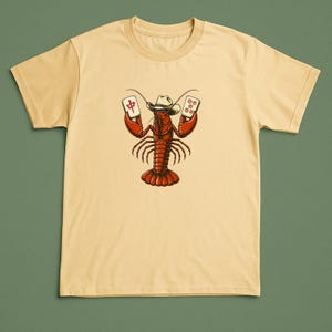 May include: A light yellow short-sleeve t-shirt featuring a graphic illustration of a red lobster wearing a white cowboy hat. The lobster is holding two mahjong tiles in its claws, one with a red Chinese character and one with five red circles.