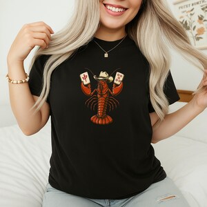 May include: A black t-shirt featuring a graphic illustration of a red lobster wearing a cowboy hat and holding two mahjong tiles. The lobster is centered on the chest, with one tile displaying a red Chinese character and the other showing five dots.