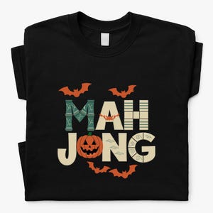 May include: Black t-shirt with a Halloween-themed design. The word "MAH JONG" is spelled out in a decorative font, with a pumpkin and bats incorporated into the design. The shirt is made of a soft material.