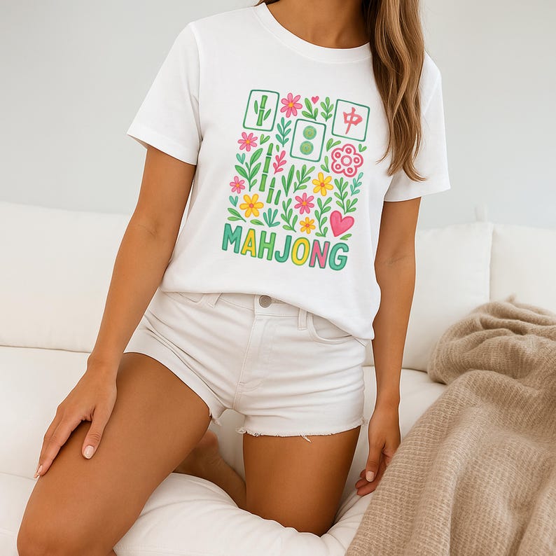 May include: White t-shirt with a colorful Mahjong design. The graphic includes floral elements, Mahjong tiles, and the word "MAHJONG" in green. The shirt is a crew neck style.