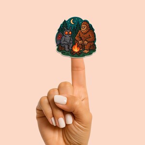 Puede incluir: Un t&iacute;tere de dedo con una ilustraci&oacute;n de dibujos animados de un Bigfoot y una polilla sentados junto a una fogata bajo una luna creciente. El t&iacute;tere est&aacute; hecho de vinilo y tiene un esquema de color verde, marr&oacute;n y naranja.