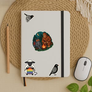 Puede incluir: Un conjunto de cinco pegatinas con diferentes ilustraciones. Las pegatinas incluyen un p&aacute;jaro negro, una polilla en blanco y negro, una criatura parecida a un pie grande, un p&aacute;jaro negro con un dise&ntilde;o de mandala y una oveja tejiendo una bufanda arco&iacute;ris.
