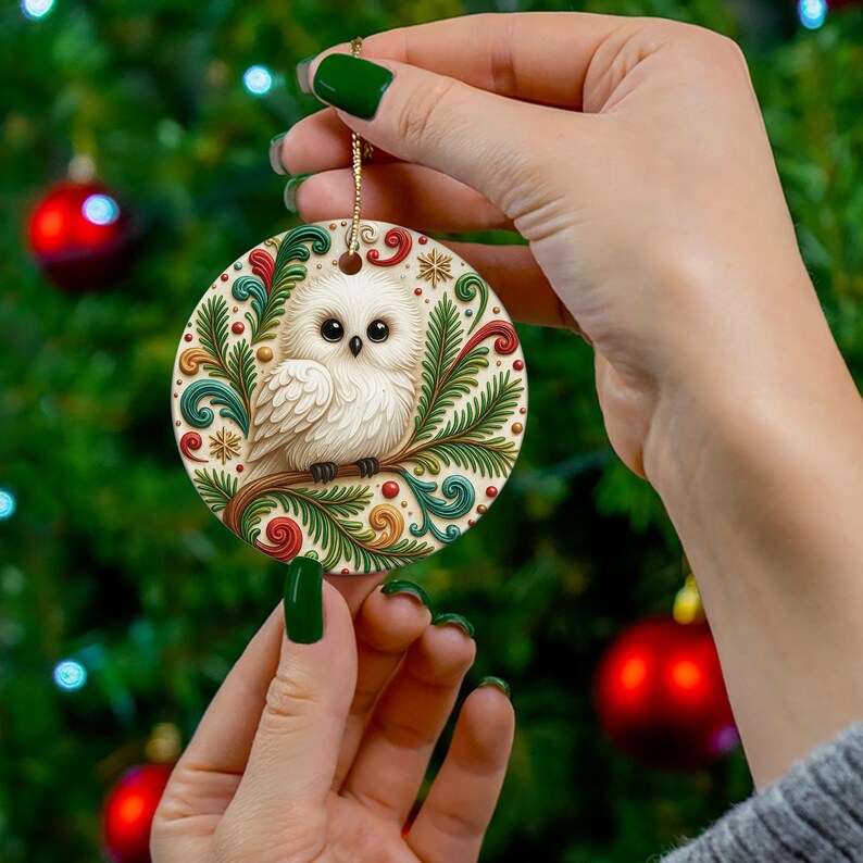 May include: A round Christmas ornament featuring a white owl perched on a branch, surrounded by green and red decorative elements. The ornament is held by a person in front of a Christmas tree.