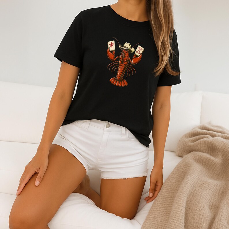 May include: A black short-sleeved t-shirt featuring a graphic print of a red lobster wearing a cowboy hat and holding two mahjong tiles. The lobster is centered on the chest area of the shirt, which is paired with white denim shorts.