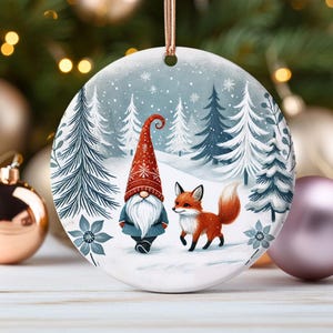 May include: A round Christmas ornament with a white background and a winter scene. A gnome with a red hat and a white beard stands next to a red fox in a snowy forest.