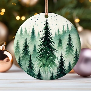 May include: A round ceramic ornament with a watercolor painting of a snowy forest. The trees are green and the background is white with small white dots.