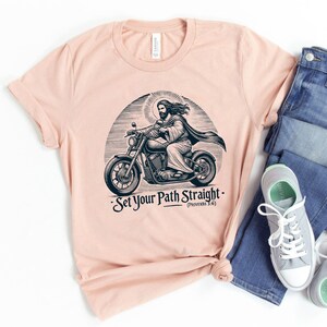 Puede incluir: Una camiseta de color melocot&oacute;n con una ilustraci&oacute;n en blanco y negro de Jes&uacute;s conduciendo una motocicleta. El texto "Set Your Path Straight - (Proverbs 3:6)" est&aacute; impreso debajo de la imagen.