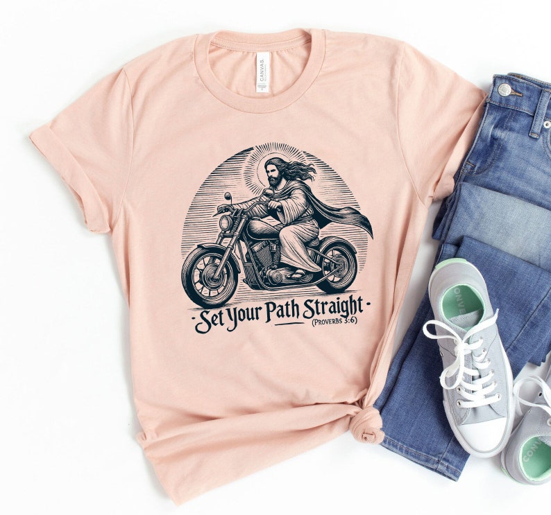 Puede incluir: Una camiseta de color melocot&oacute;n con una ilustraci&oacute;n en blanco y negro de Jes&uacute;s conduciendo una motocicleta. El texto "Set Your Path Straight - (Proverbs 3:6)" est&aacute; impreso debajo de la imagen.