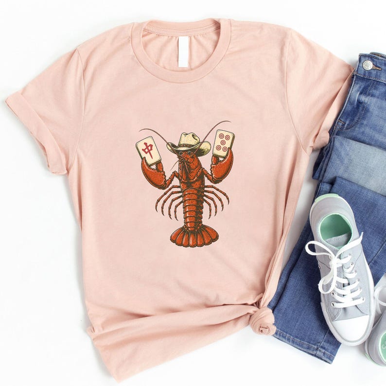 May include: A light pink short-sleeve t-shirt featuring a graphic illustration of a red lobster wearing a beige cowboy hat. The lobster is holding two mahjong tiles, one with a red Chinese character and the other with five red circles.