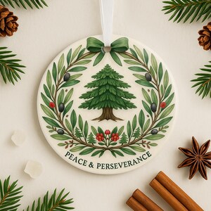 May include: A round, white ceramic ornament with a green wreath design featuring a green pine tree in the center. The wreath is decorated with red berries and black olives. The ornament has a white ribbon and the text "Peace & Perseverance" at the bottom.