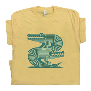 Peut inclure: Un t-shirt jaune avec un motif bleu turquoise de deux alligators stylis&eacute;s. Les alligators sont tourn&eacute;s vers l'avant, la gueule ouverte, et leur corps est compos&eacute; de lignes.