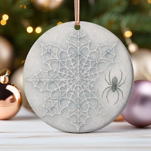 May include: A round, light gray Christmas ornament with a snowflake design and a spider illustration. The snowflake is detailed with a web-like pattern and small, silver-colored embellishments. A gray spider is on the right side of the ornament, and a gold ribbon is attached for hanging.