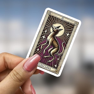 May include: A sticker featuring a tarot card with a purple background and gold accents. The card depicts a creature with a skeletal appearance, with the text "VII  CHUPACABRA" at the bottom.