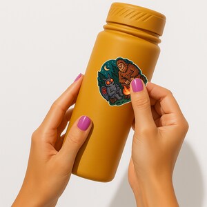 Puede incluir: Una botella de agua amarilla con una pegatina de un Bigfoot y una polilla sentados junto a una fogata. La pegatina tiene un esquema de color azul, verde y naranja.
