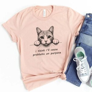 May include: A light pink t-shirt with a black and white illustration of a cat peeking over the top. The text "i think i'll cause problems on purpose" is printed below the cat.