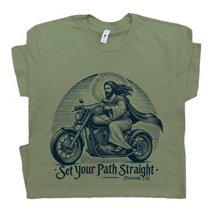Puede incluir: Una camiseta verde con un gr&aacute;fico de Jes&uacute;s conduciendo una motocicleta. El gr&aacute;fico es en blanco y negro y tiene un aspecto vintage. El texto en la camiseta dice "Set Your Path Straight - (Proverbs 3:6)"