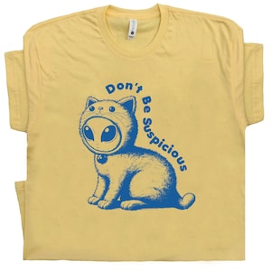 May include: A yellow t-shirt with a blue cartoon cat wearing an alien mask. The text "Don't Be Suspicious" is written in a circle around the cat.