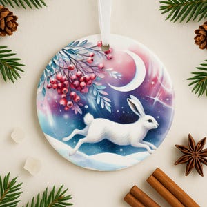 May include: A round ceramic ornament featuring a white rabbit running across a snowy landscape under a crescent moon and starry sky. The ornament has a white ribbon for hanging and is decorated with red berries and foliage.