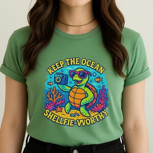May include: Green t-shirt featuring a cartoon turtle wearing sunglasses and holding a camera. The graphic includes the text "KEEP THE OCEAN SHELLFIE-WORTHY" in a colorful design with coral, fish, and a starfish. The shirt is made of a soft material.