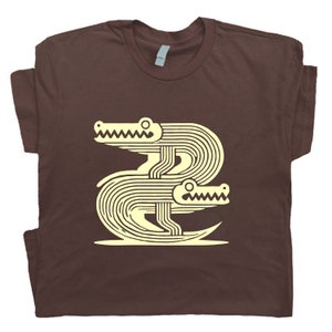 Peut inclure: Un t-shirt marron avec un motif graphique de deux alligators stylis&eacute;s en jaune clair.