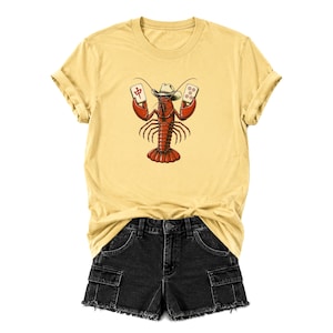 May include: A pale yellow t-shirt featuring a graphic of a red lobster wearing a cowboy hat and holding two mahjong tiles. The lobster is centered on the chest. Below the t-shirt are black denim shorts with a frayed hem and cargo pockets.