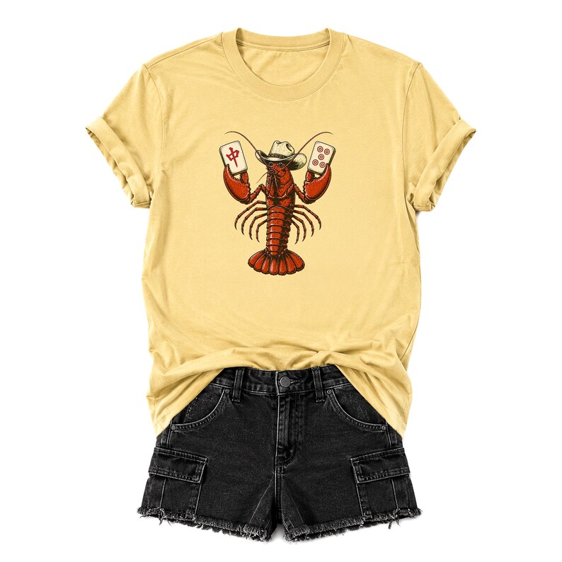 May include: A pale yellow t-shirt featuring a graphic of a red lobster wearing a cowboy hat and holding two mahjong tiles. The lobster is centered on the chest. Below the t-shirt are black denim shorts with a frayed hem and cargo pockets.