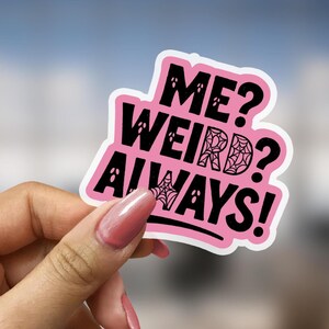 May include: A pink sticker with black text that reads "ME? WEIRD? ALWAYS!" The sticker has a white border and features spiderwebs and ghost-like figures. The sticker is held by a person.