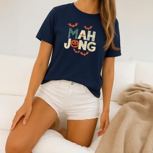 May include: Navy blue t-shirt with the word "MAH JONG" in a Halloween-themed design. The design includes a jack-o'-lantern and bats. The shirt is paired with white shorts.