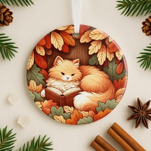 May include: Round ceramic ornament featuring a fluffy orange cat reading a book under a tree with autumn leaves. The ornament has a white ribbon for hanging. The design includes shades of orange, brown, and green, evoking a cozy, seasonal theme.