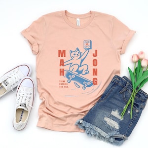 May include: A peach colored t-shirt with a graphic of a cat riding a skateboard. The cat is holding a mahjong tile. The text "Mahjong" is written vertically on the left side of the graphic and the text "Think outside the tile" is written below the graphic.