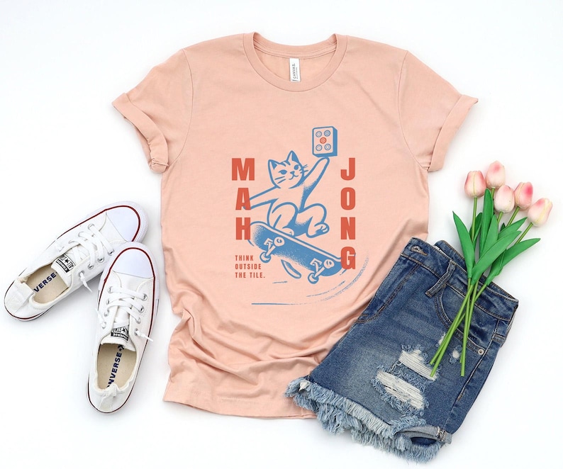 May include: A peach colored t-shirt with a graphic of a cat riding a skateboard. The cat is holding a mahjong tile. The text "Mahjong" is written vertically on the left side of the graphic and the text "Think outside the tile" is written below the graphic.