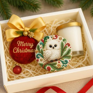 May include: A gift box containing a red ornament with "Merry Christmas" text, a gold bow, a white owl ornament, a small red ornament, and a white candle with a gold lid. The box is filled with straw.