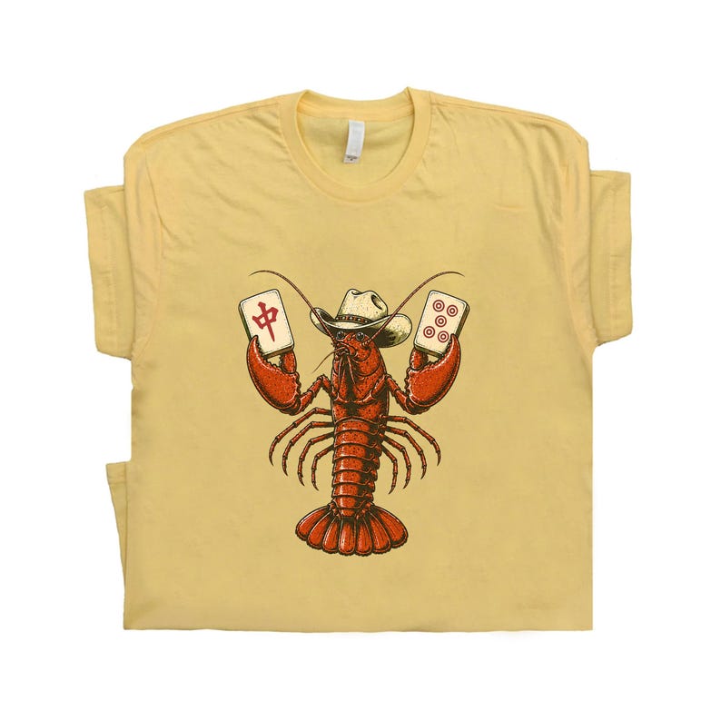May include: A pale yellow t-shirt featuring a graphic illustration of a red lobster wearing a white cowboy hat. The lobster is holding two mahjong tiles, one with the red Chinese character for middle and one with five red circles.