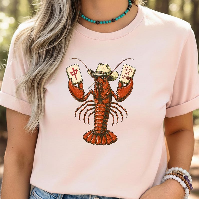 May include: A light pink t-shirt featuring a graphic illustration of a red lobster wearing a cowboy hat. The lobster is holding a mahjong tile with the Chinese character for middle in one claw and a mahjong tile with five circles in the other.