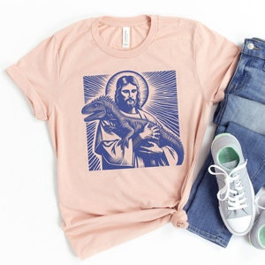 May include: A light pink t-shirt with a blue graphic print of Jesus holding a lizard.