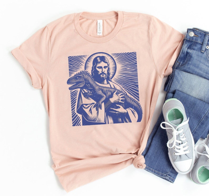 May include: A light pink t-shirt with a blue graphic print of Jesus holding a lizard.