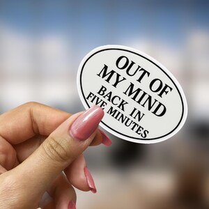 May include: An oval sticker with a white background and a black border. The text "OUT OF MY MIND BACK IN FIVE MINUTES" is printed in black, centered on the sticker. The sticker is held by a hand with pink nail polish.