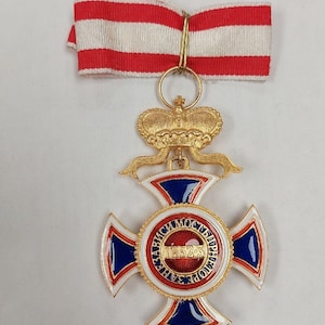 Puede incluir: Una medalla de oro con una cruz de esmalte rojo, blanco y azul. El centro de la cruz presenta un círculo rojo con el texto "1852-3" en oro. La medalla está suspendida de una cinta roja y blanca.