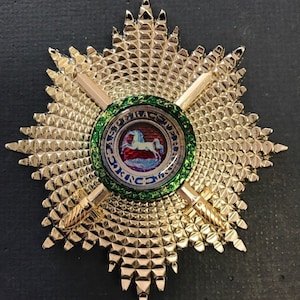 May include: A gold-coloured star-shaped emblem with a central circular design. The emblem features a detailed pattern of small, raised squares and a green and gold border. The centre design includes a white horse and text.