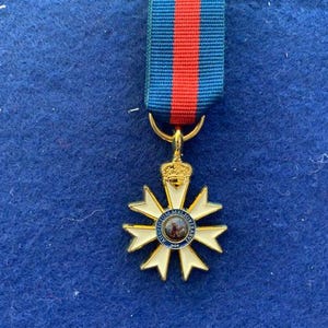 Op de afbeelding: Een gouden en witte medaille met een blauw en wit centrum met een portret. De medaille hangt aan een blauw en rood lint.