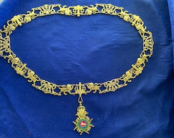 May include: A gold chain with lion and crown motifs, ending in a pendant with a red and green emblem and a gold star.
