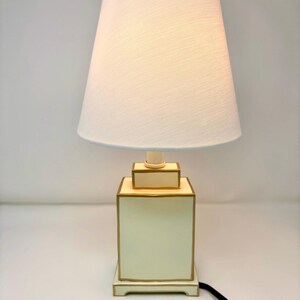 May include: A cream-colored table lamp with a white lampshade. The lamp base is a square shape with gold trim. The lampshade is a light cream color with a light blue trim. A black cord extends from the base.