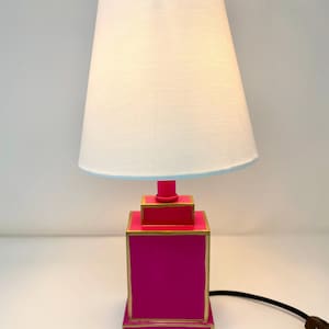 May include: A vibrant pink table lamp with a white lampshade and a light blue trim. The lamp base is a stacked square design with gold trim. A black cord and brown plug are visible.