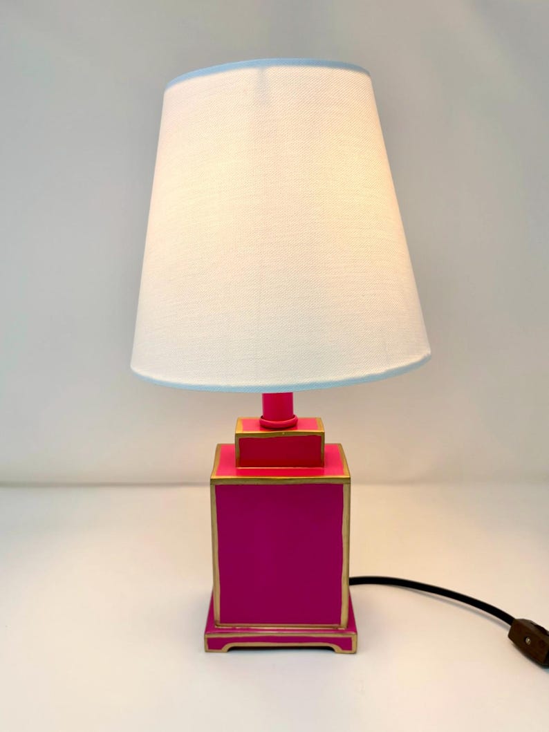 May include: A vibrant pink table lamp with a white lampshade and a light blue trim. The lamp base is a stacked square design with gold trim. A black cord and brown plug are visible.