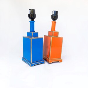 May include: Two decorative table lamps. One is bright blue, the other is orange. Both have a rectangular base with gold trim, a cylindrical neck, and a black lamp socket. The lamps are set against a white background.