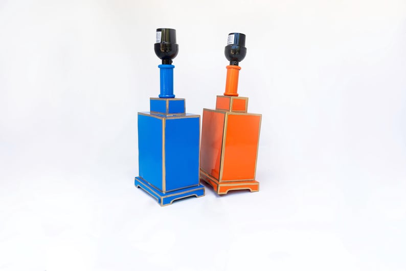 May include: Two decorative table lamps. One is bright blue, the other is orange. Both have a rectangular base with gold trim, a cylindrical neck, and a black lamp socket. The lamps are set against a white background.