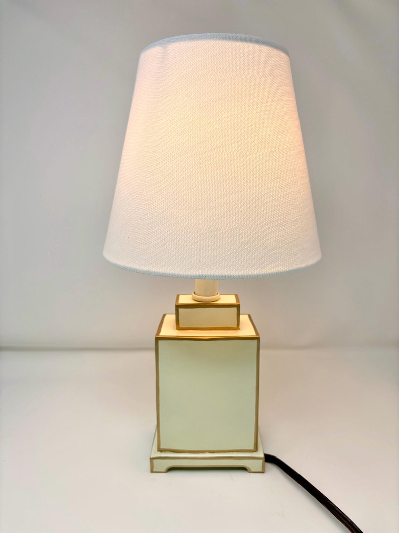 May include: A cream-colored table lamp with a white lampshade. The lamp base is a square shape with gold trim. The lampshade is a light cream color with a light blue trim. A black cord extends from the base.