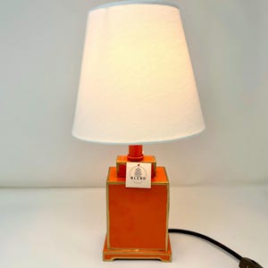 May include: An orange table lamp with a white lampshade and a light blue trim. The lamp has a rectangular base with gold trim and a matching orange stem. A tag with the text "BLEND" is attached to the lamp.